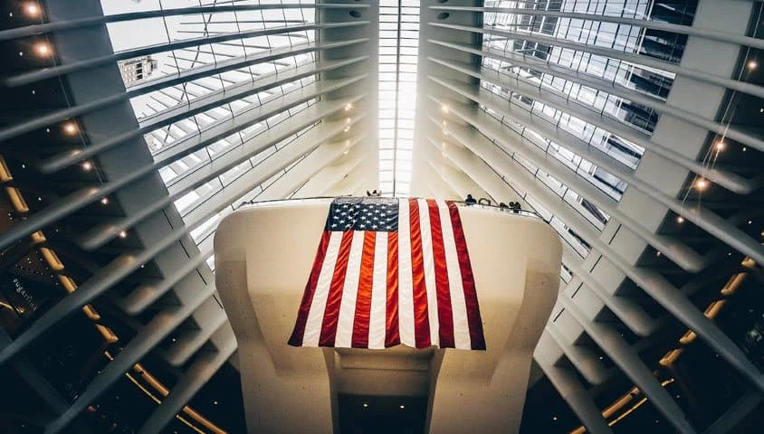 A large American flag hangs inside a modern, bright building with white beams and a skylight ceiling, viewed from below. People stand on a balcony above the flag.