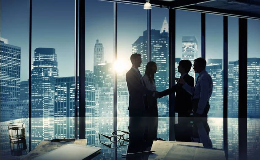 Four businesspeople stand and shake hands in a modern office with large windows, city skyscrapers illuminated at dusk in the background. Papers, a pen, and glasses are on a reflective table in the foreground.