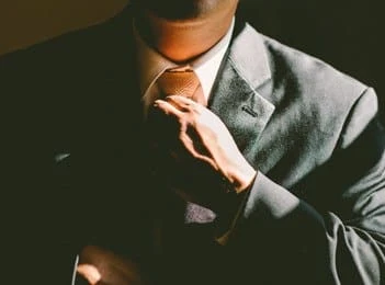 A person in a suit adjusts their tie, partially illuminated by light, with their face mostly in shadow.