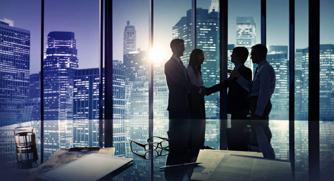 Four businesspeople stand and talk in front of large office windows with a city skyline in the background; papers, glasses, and a glass of water are on a reflective table in the foreground.