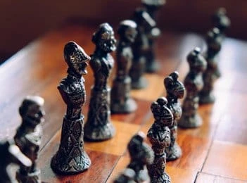 Close-up of intricately designed metal chess pieces arranged on a wooden chessboard, with the focus on the front pieces and the background pieces blurred.
