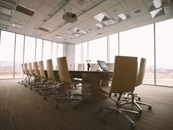 A modern conference room with a long table surrounded by empty swivel chairs, large floor-to-ceiling windows, and natural light filling the space.