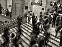 Black and white photo of a busy indoor staircase with many people walking up and down, creating a sense of motion and crowd in a public building.