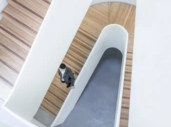 A person walks on wooden stairs between white curved walls in a modern, minimalist interior, viewed from above.