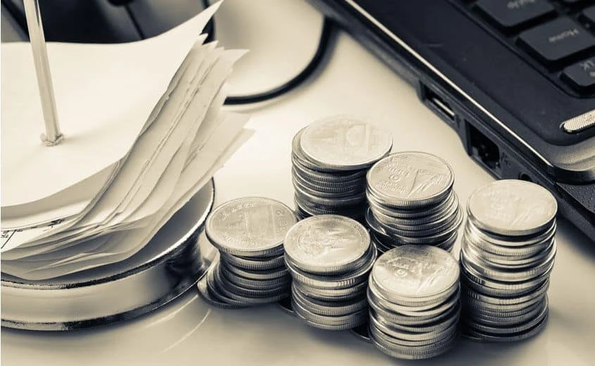 Stacks of coins are arranged next to a pile of receipts on a metal spindle and a laptop keyboard on a white desk. The image is in black and white.