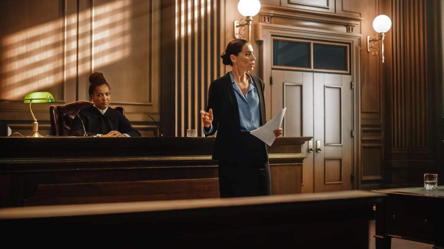 A lawyer stands and speaks in a courtroom, holding documents, while a judge sits behind the bench listening attentively. The room has wooden walls, large doors, and round lights.