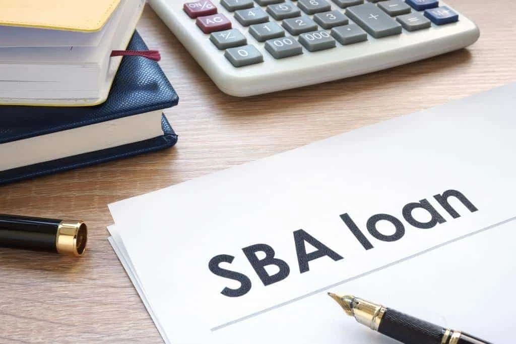A document labeled "SBA loan" on a desk next to a fountain pen, stacked notebooks, and a calculator.