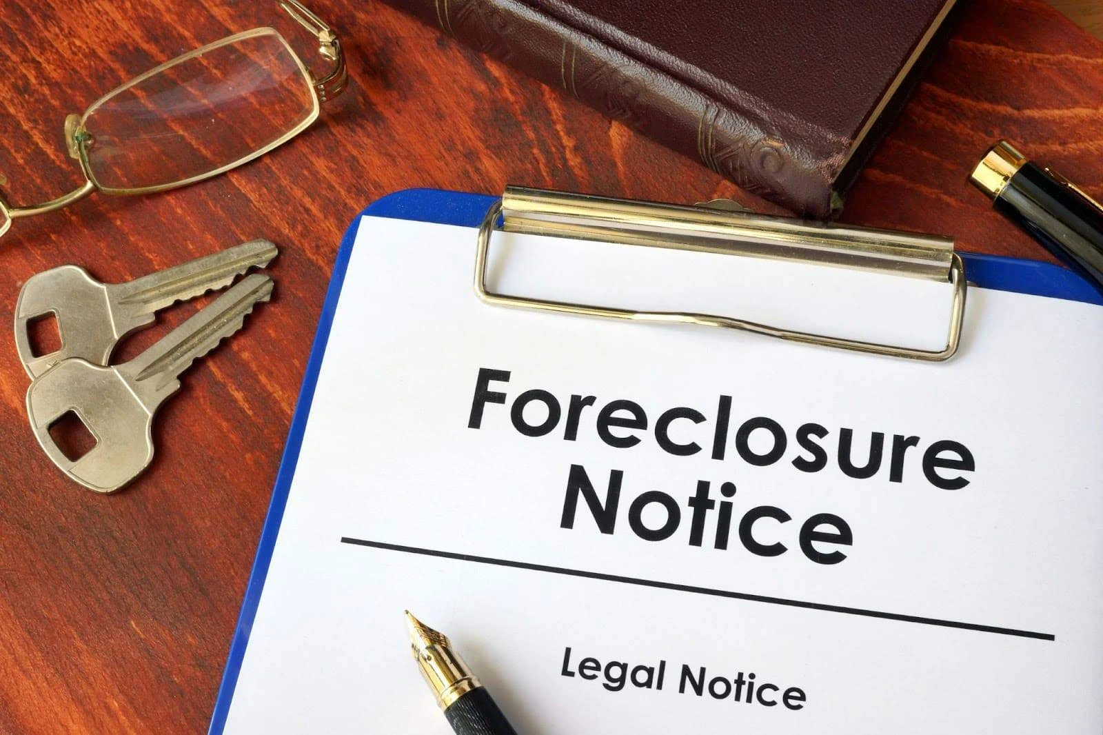 A clipboard with a document titled "Foreclosure Notice" and "Legal Notice" on a wooden desk, next to a pair of keys, a pen, eyeglasses, and a closed book.
