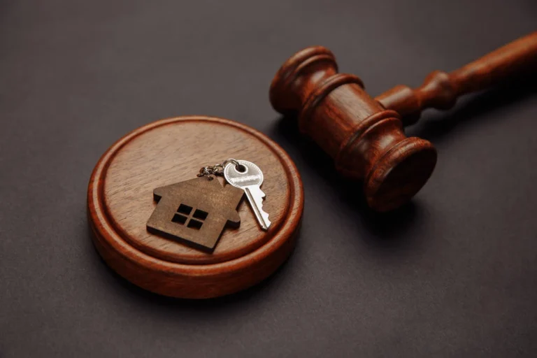 A wooden judge’s gavel rests beside a round base with a house-shaped keychain and a silver key on it, symbolizing real estate law or property auctions.