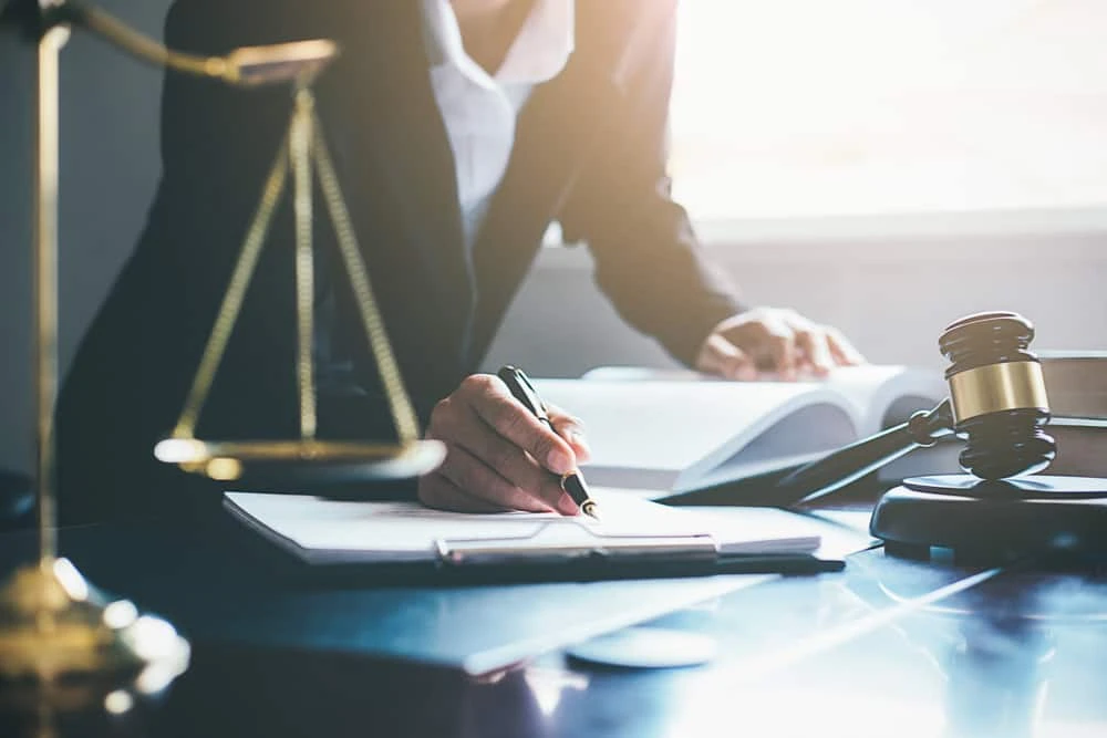 A person in a suit writes in a notebook at a desk with a gavel, scales of justice, and legal books, suggesting a legal or courtroom setting. Sunlight streams in through a window in the background.