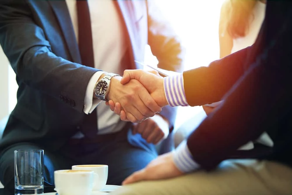 Two people in business attire shaking hands in a well-lit room, symbolizing an agreement or partnership. A third person, perhaps a bankruptcy lawyer, is partially visible in the background. A table with cups and glasses is in the foreground, setting the scene for potential legal discussions.