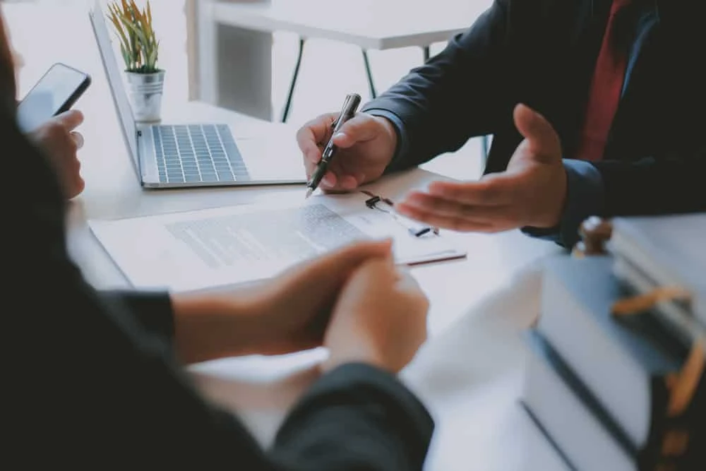 Two people in business attire are seated at a desk with a laptop and documents. One, likely a bankruptcy lawyer, holds a pen, gesturing towards the paperwork, while the other listens attentively. A small plant and stack of books are on the table.