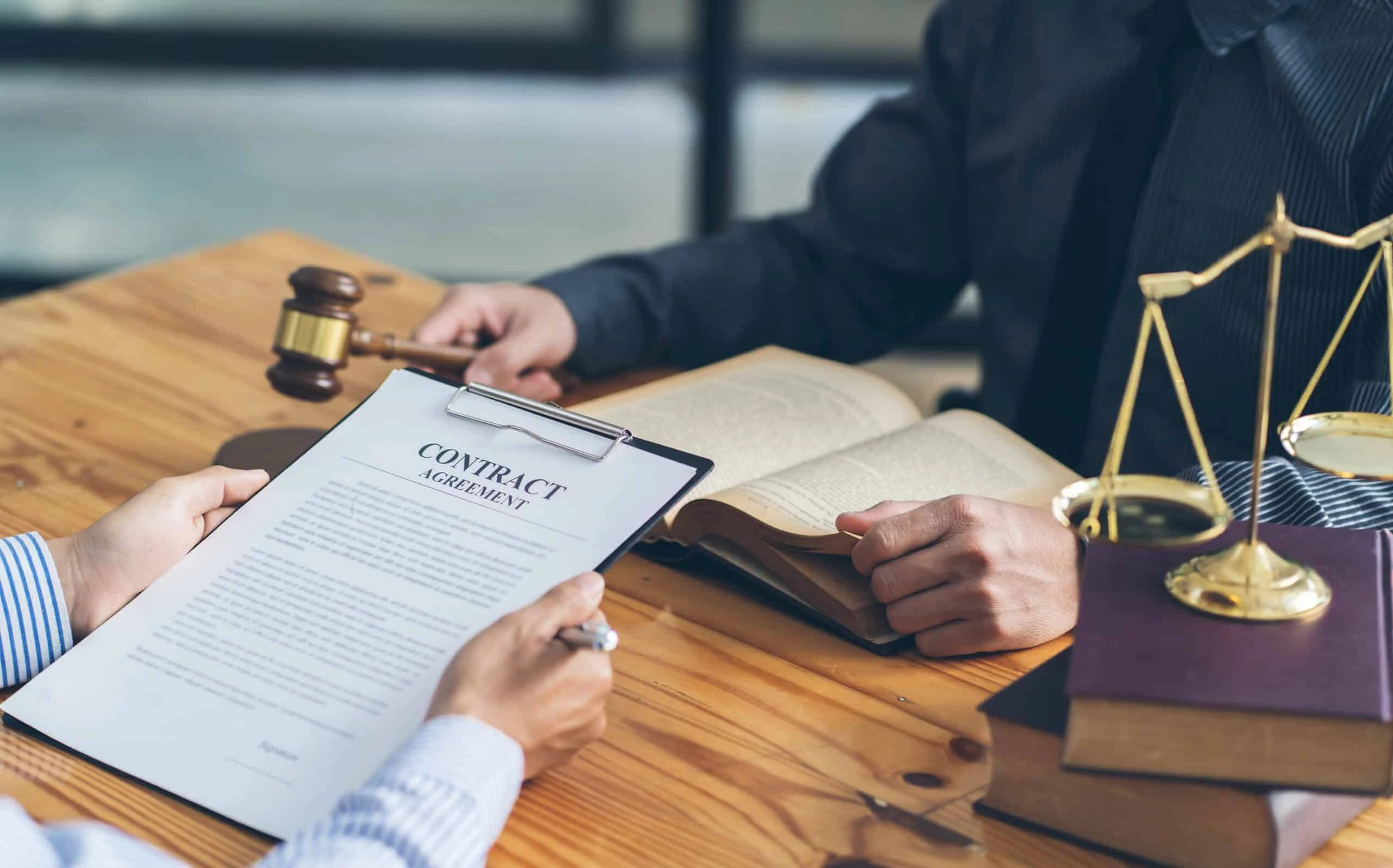 Two people sit at a wooden table, with one holding a contract on a clipboard and pen, while the other is next to an open book. A gavel and scales of justice suggest legal work, possibly involving a bankruptcy lawyer, as they delve into financial matters.