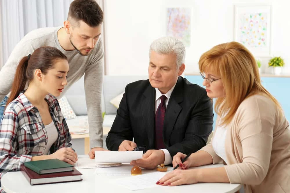 Four people sit around a table cluttered with documents. An older man in a suit, possibly a foreclosure attorney, points at papers while two women and a younger man focus intently on the real estate law discussion. The office setting, filled with books and papers, hints at civil litigation matters underway.