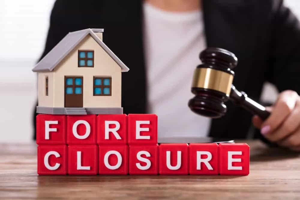 A foreclosure attorney in a suit holds a gavel, seated at a table. Before them, a toy house rests on red blocks spelling "FORECLOSURE," symbolizing the gravity of civil litigation in real estate disputes.