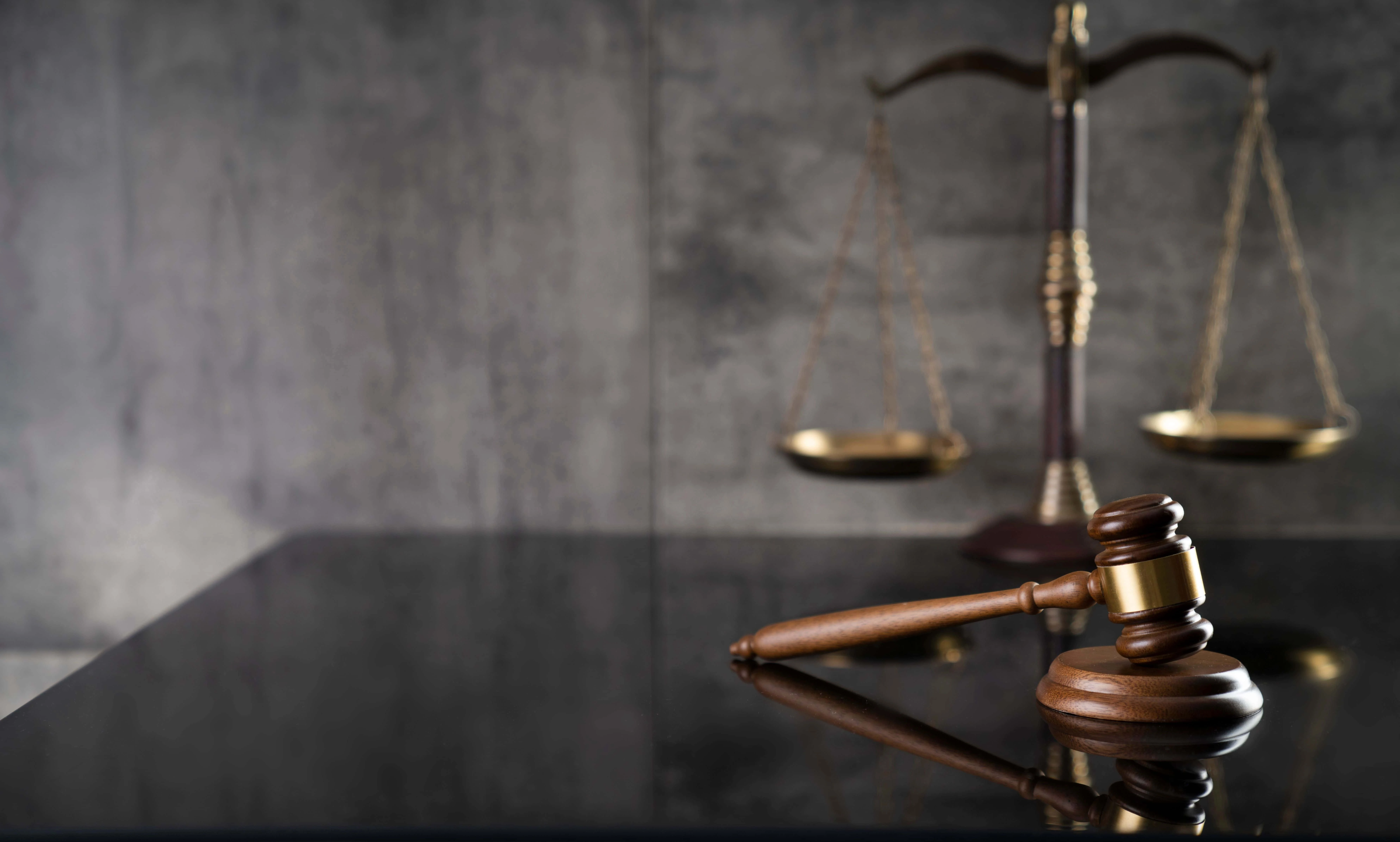 A wooden judge's gavel rests on a reflective black surface in the foreground. In the background, a balance scale stands against a gray textured wall, symbolizing law and justice, much like the tools of a foreclosure attorney navigating complex litigation.