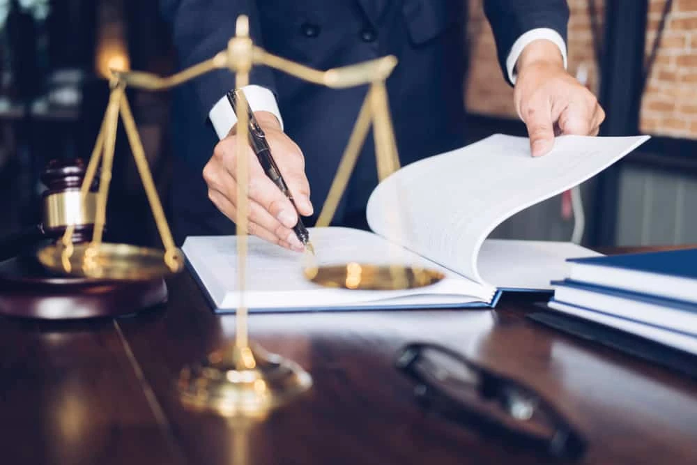 A person in formal attire stands behind a desk, signing a document. Scales of justice, a gavel, and a pair of glasses lie on the desk, suggesting a legal environment typical of litigation or real estate law. The background is slightly blurred, featuring a brick wall.