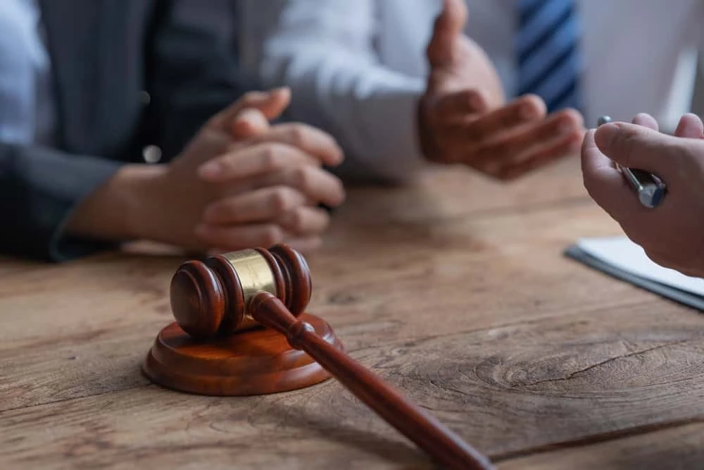 Close-up of a wooden gavel on a table, with three people discussing in the background. Two have hands clasped while another gestures openly. A notebook and pen lie nearby, hinting at a foreclosure attorney's role in real estate law or civil litigation matters being deliberated.