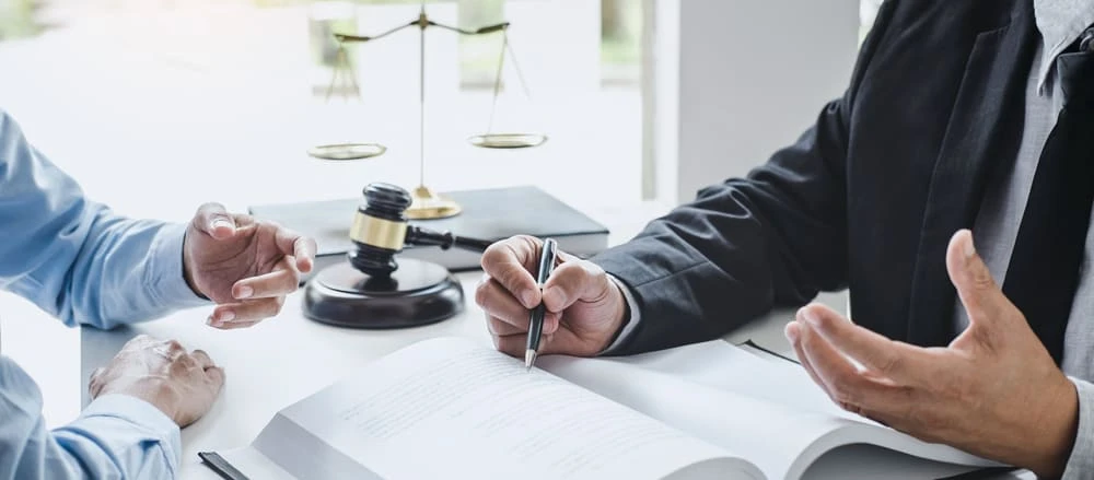 Two people are seated at a table, engaged in discussion. Legal items like a gavel, scales, and an open document are visible, suggesting a professional meeting with a focus on litigation. One person gestures while the other holds a pen, ready to write or sign.