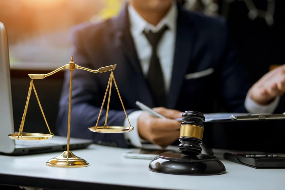 A foreclosure attorney in a suit sits at a desk with a scale of justice and a gavel prominently displayed. They are writing on a document, with a laptop nearby. The room is softly lit, suggesting a legal or professional setting focused on real estate law.