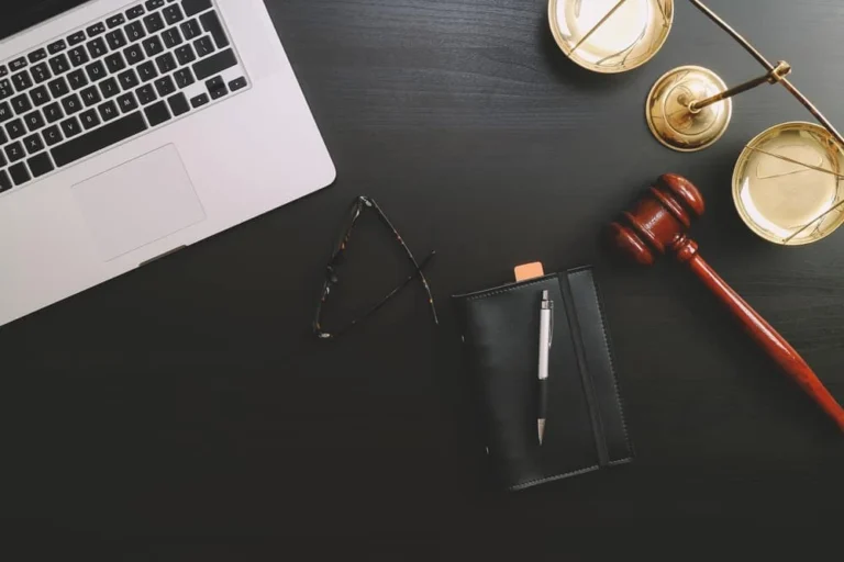 A laptop, eyeglasses, and a notebook with a pen lie on a dark desk. A wooden gavel and golden scales of justice hint at a civil litigation setting, emphasizing the professional atmosphere.