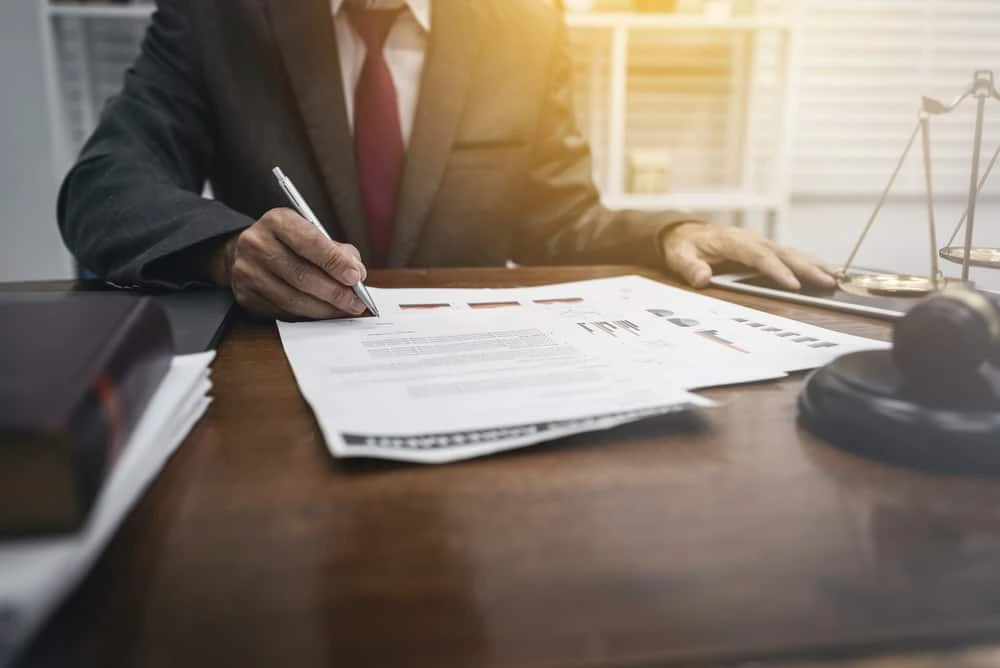 A person in a suit sits at a desk, meticulously reviewing documents. The table is covered with papers, a book on real estate law, and a set of scales. Soft sunlight filters through the window, illuminating the scene and suggesting a professional legal setting.