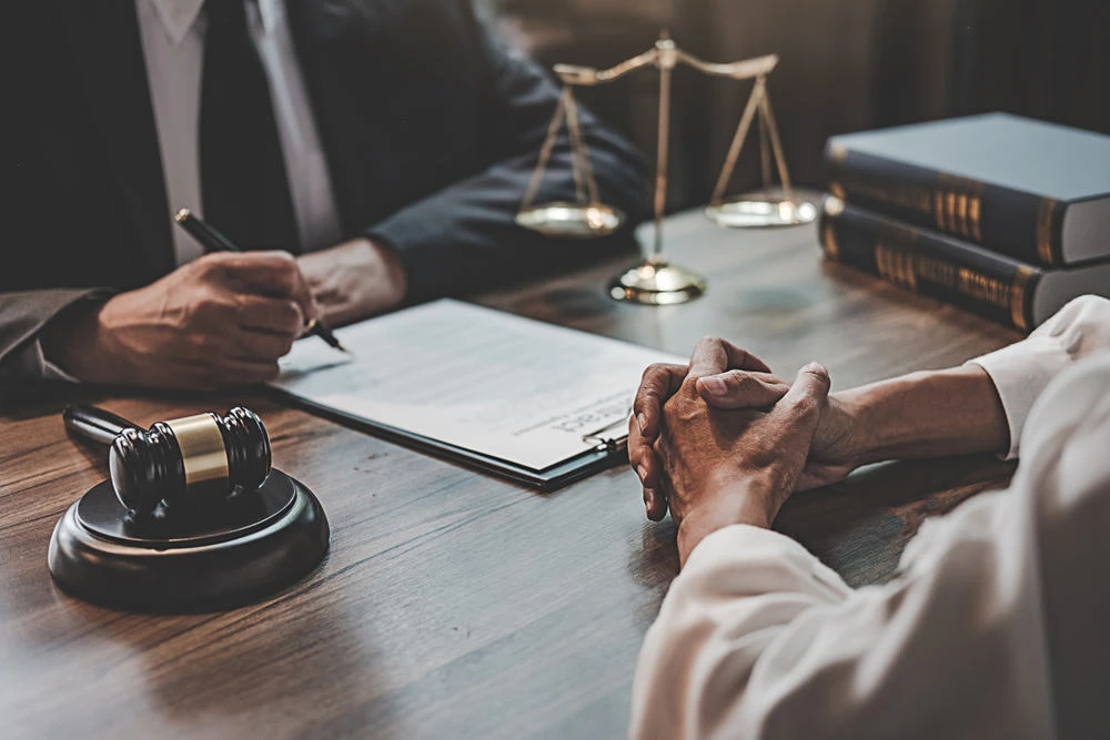 Two people sit across a wooden desk. One is signing a document with a pen, while the other has hands clasped, indicative of their role as a foreclosure attorney. A gavel, scales of justice, and law books hint at the realm of real estate law in this professional setting.