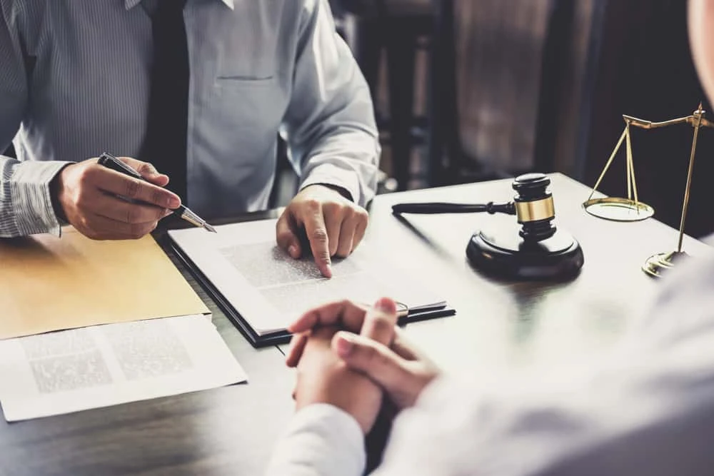 Two people are seated at a table with legal documents. One, possibly a bankruptcy lawyer, points to a document with a pen while the other listens intently. A judge's gavel and balance scale are on the table, suggesting a severe litigation or consultation setting.