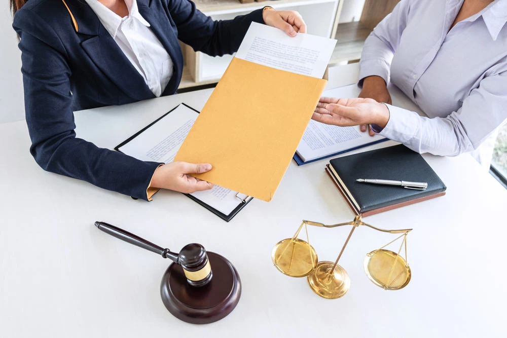 Two individuals sit at a table strewn with legal documents, a gavel, and a brass balance scale. One hands an envelope marked "foreclosure" to the other. Both are in business attire, with a notebook and pen lying next to them, capturing the gravity of their discussion.