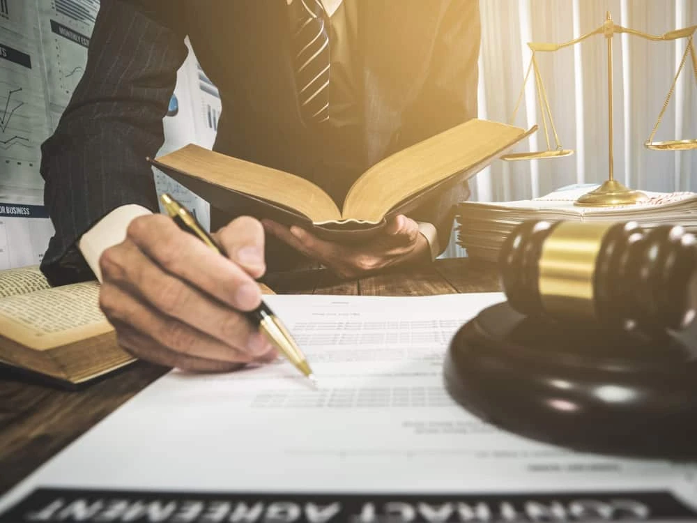 A person in a suit holds an open book and writes on a document at a desk. A gavel, scales of justice, and stacks of papers denote a litigation setting. The scene is well-lit, suggesting a civil litigation or professional environment.