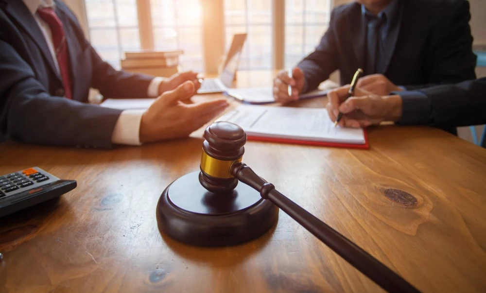 A gavel rests on a wooden table in the foreground. In the background, two people in suits are seated, one gesturing while the other writes on a document related to civil litigation. A calculator and books are also visible on the table.