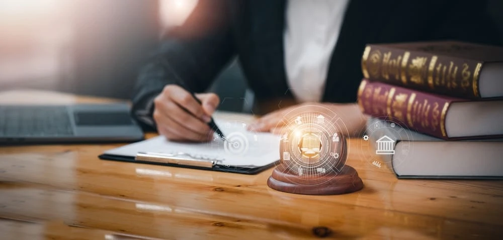 A person in a suit writes on a clipboard at a wooden desk. A laptop and stacked law books are nearby. A digital gavel icon with symbols like scales and a courthouse floats above, emphasizing the realm of litigation in Real Estate Law.