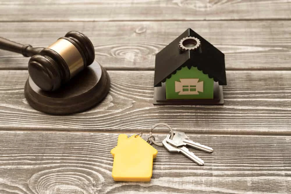 A gavel rests on a wooden surface beside a small model house with a green front and black roof, hinting at the involvement of a foreclosure attorney. In front, there are house keys attached to a yellow keychain in the shape of a house.