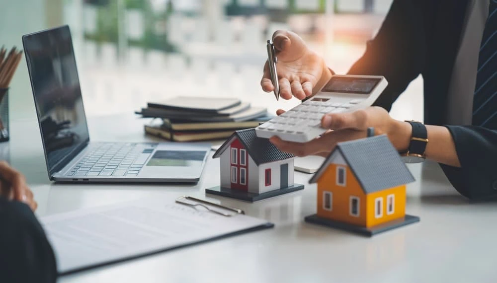 A person in business attire holds a calculator, pointing towards it with a pen, embodying the focus of a foreclosure attorney. On the desk are two small model houses, a laptop, and a clipboard with paper. The brightly lit room with large windows underscores their expertise in Real Estate Law.