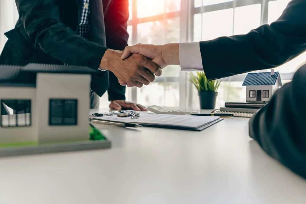 Two people shaking hands over a desk with a contract, clipboard, and model houses, as they consult on civil litigation. A plant is in the background, and sunlight streams through the large windows, giving the office a professional atmosphere.