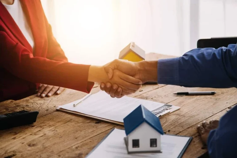 Two people are shaking hands over a wooden table with papers and a small house model on it, symbolizing an agreement rooted in Real Estate Law. One person wears a red blazer, the other a blue shirt. A pen and document are visible as sunlight streams in from the background.