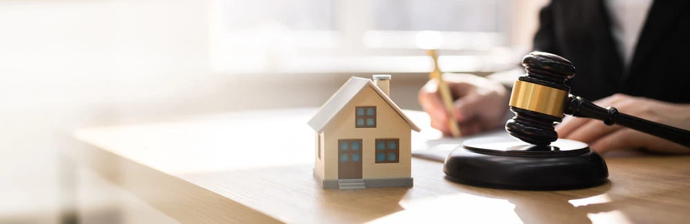 A small model house sits on a wooden table next to a judge's gavel, hinting at civil litigation in real estate. In the background, a person writes on a document, suggesting ongoing legal proceedings. Sunlight brightly illuminates the scene.