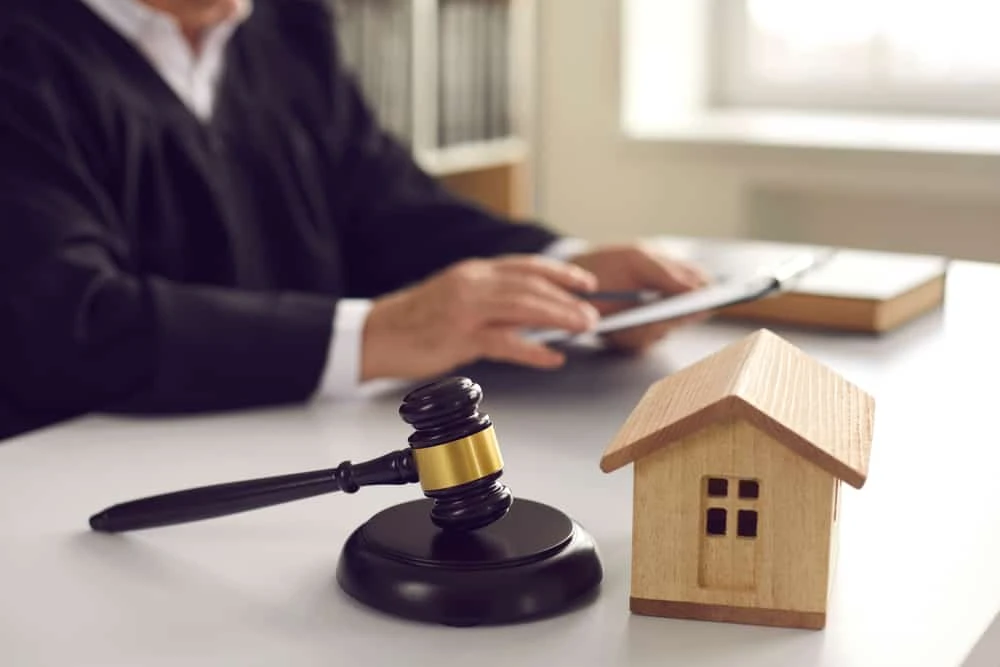 A judge in a black robe uses a tablet at a desk, likely navigating through real estate law. In the foreground, a wooden gavel rests beside a small wooden house model. Books are partially visible in the background, hinting at complex litigation matters.
