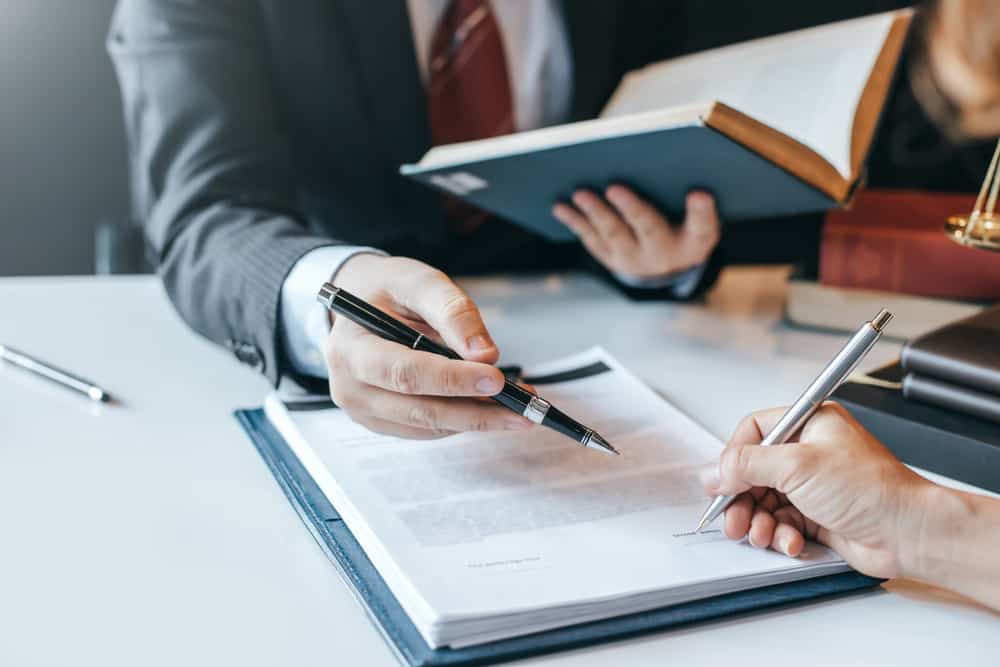 Two people in formal attire are at a desk. One holds a pen and points at a document, while the other writes. Open books and papers on civil litigation are scattered across the table, suggesting a professional setting.