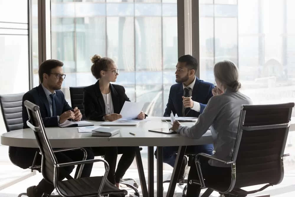Four business professionals sit around a conference table in a modern office, engaged in a discussion with documents and notebooks in front of them. Large windows reveal a cityscape in the background.