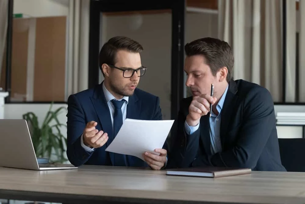Two men in business suits sit at a desk with a laptop, discussing documents. One man is holding papers and gesturing, while the other listens attentively and holds a pen.