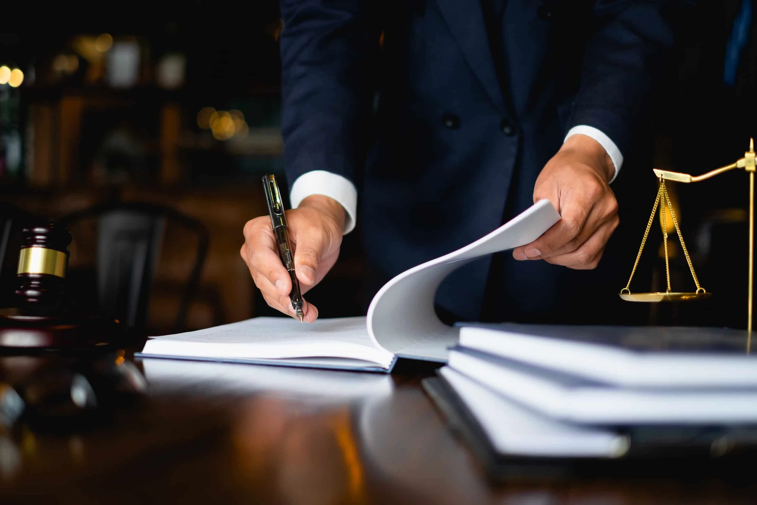 A person in a suit stands at a desk, signing a document with a pen. Nearby are a gavel, scales of justice, and stacks of books, suggesting a legal or courtroom setting.