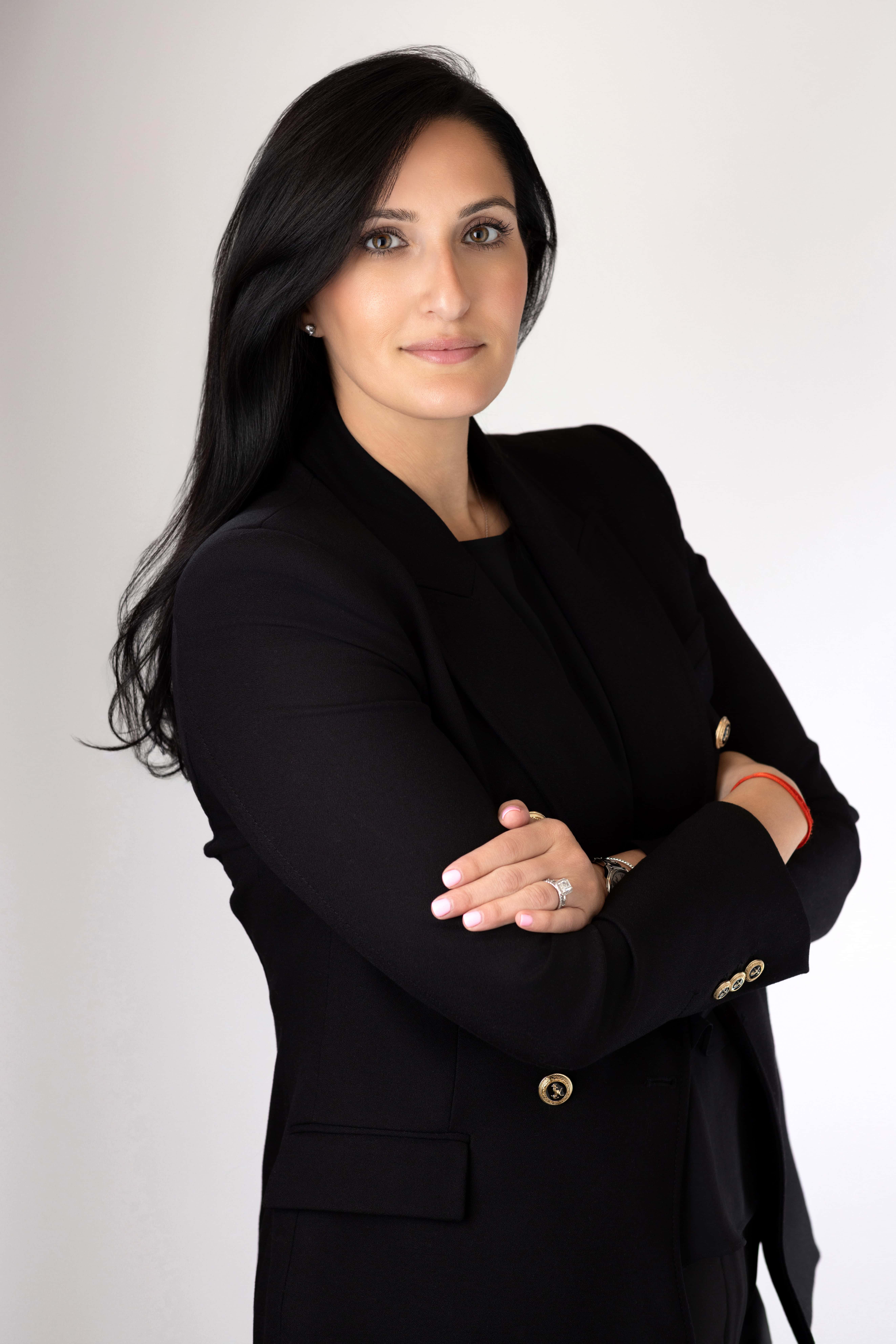 Person wearing a black suit jacket and black shirt, standing with arms crossed against a plain white background, looking directly at the camera.