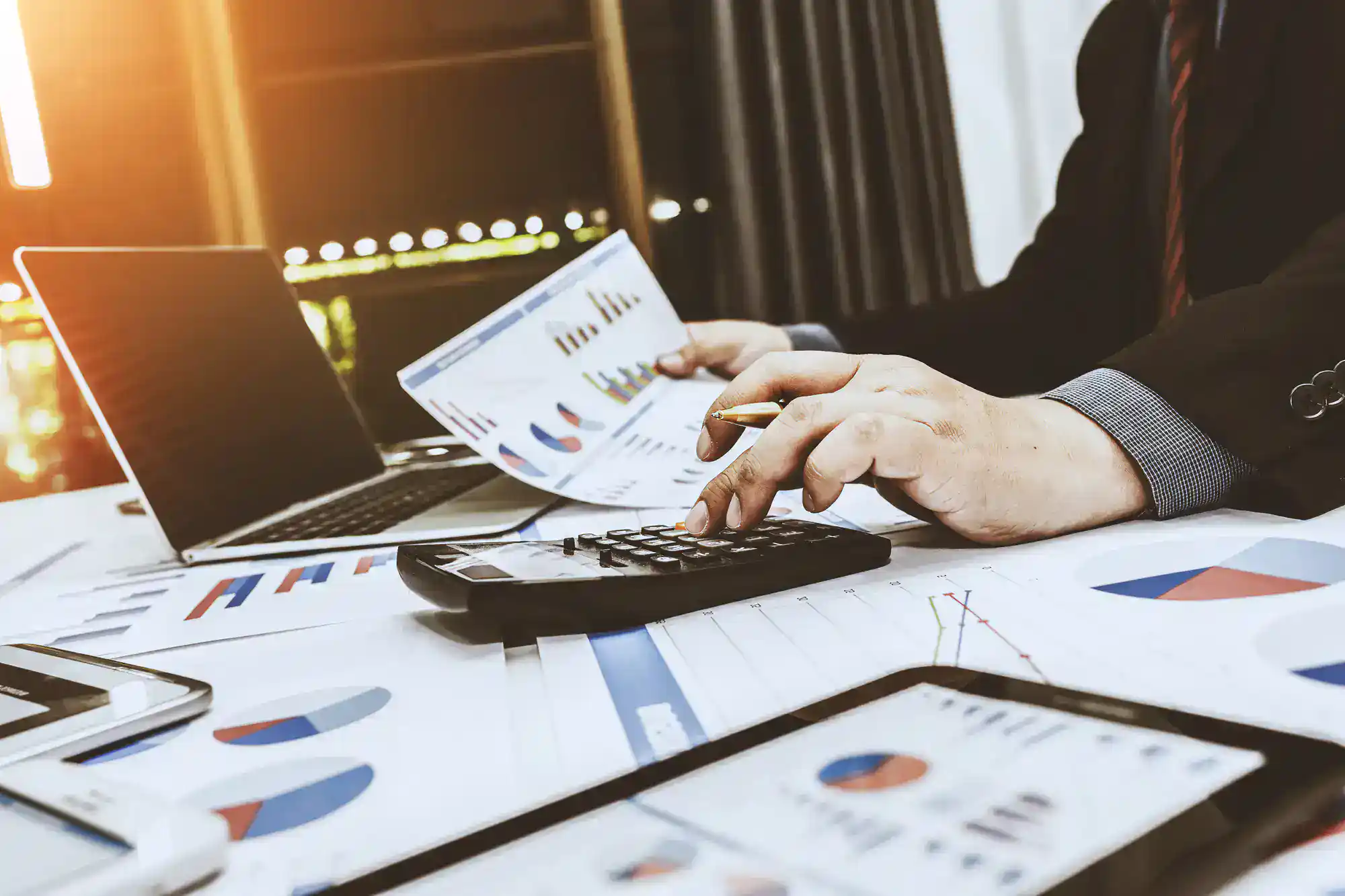 A person in business attire uses a calculator while reviewing financial charts and graphs on paper, with a laptop and documents spread out on the desk.