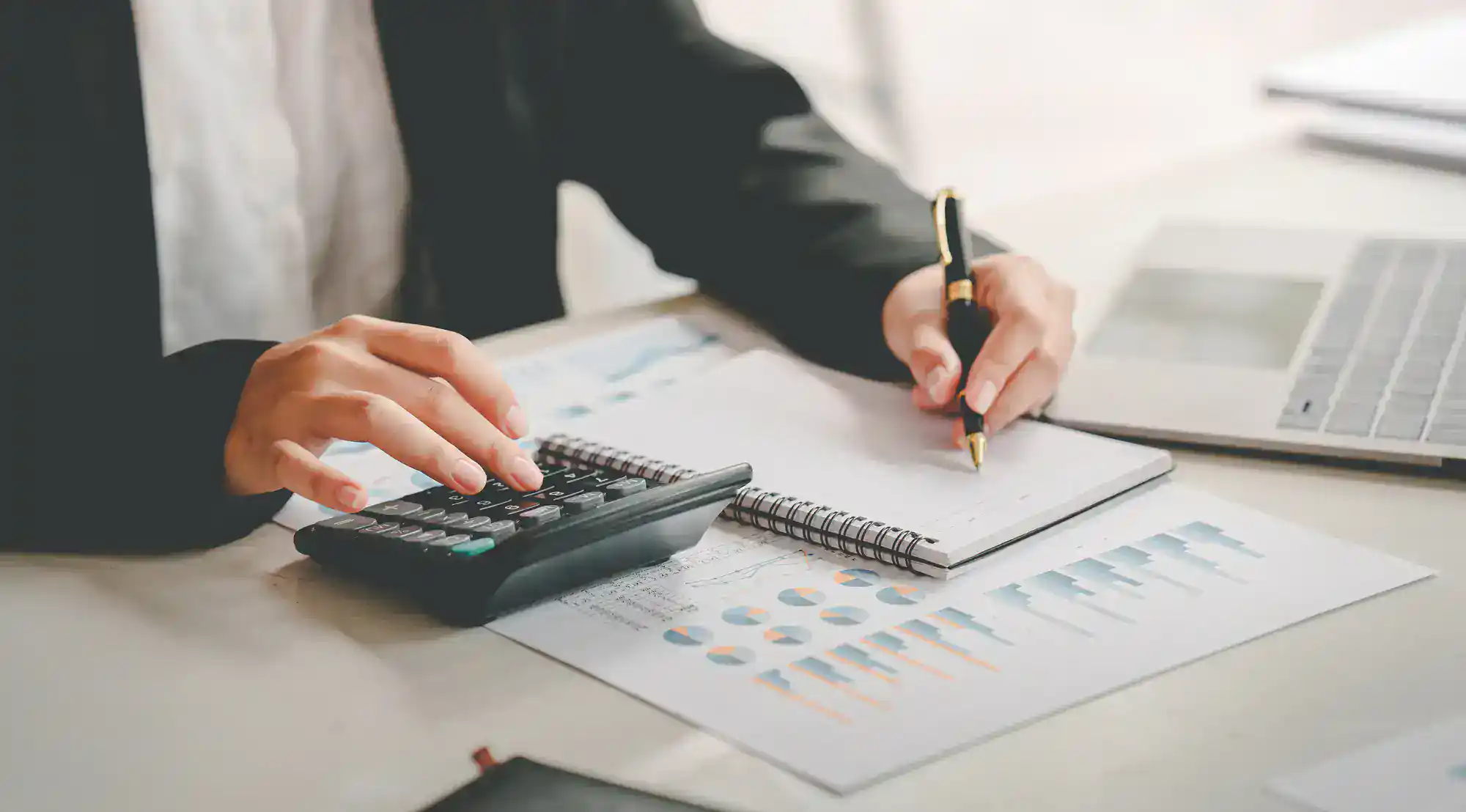 A person in business attire uses a calculator and writes in a notebook, with financial charts and a laptop on the desk, suggesting financial analysis or accounting work.