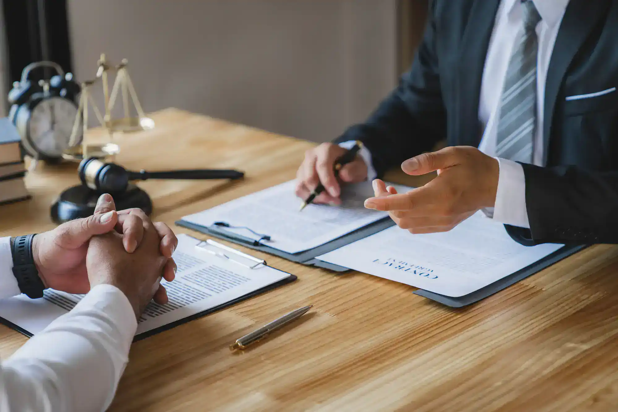 Two people in business attire sit at a desk with contract documents, a clipboard, a pen, a judge&rsquo;s gavel, and legal scales, suggesting a discussion or legal consultation.