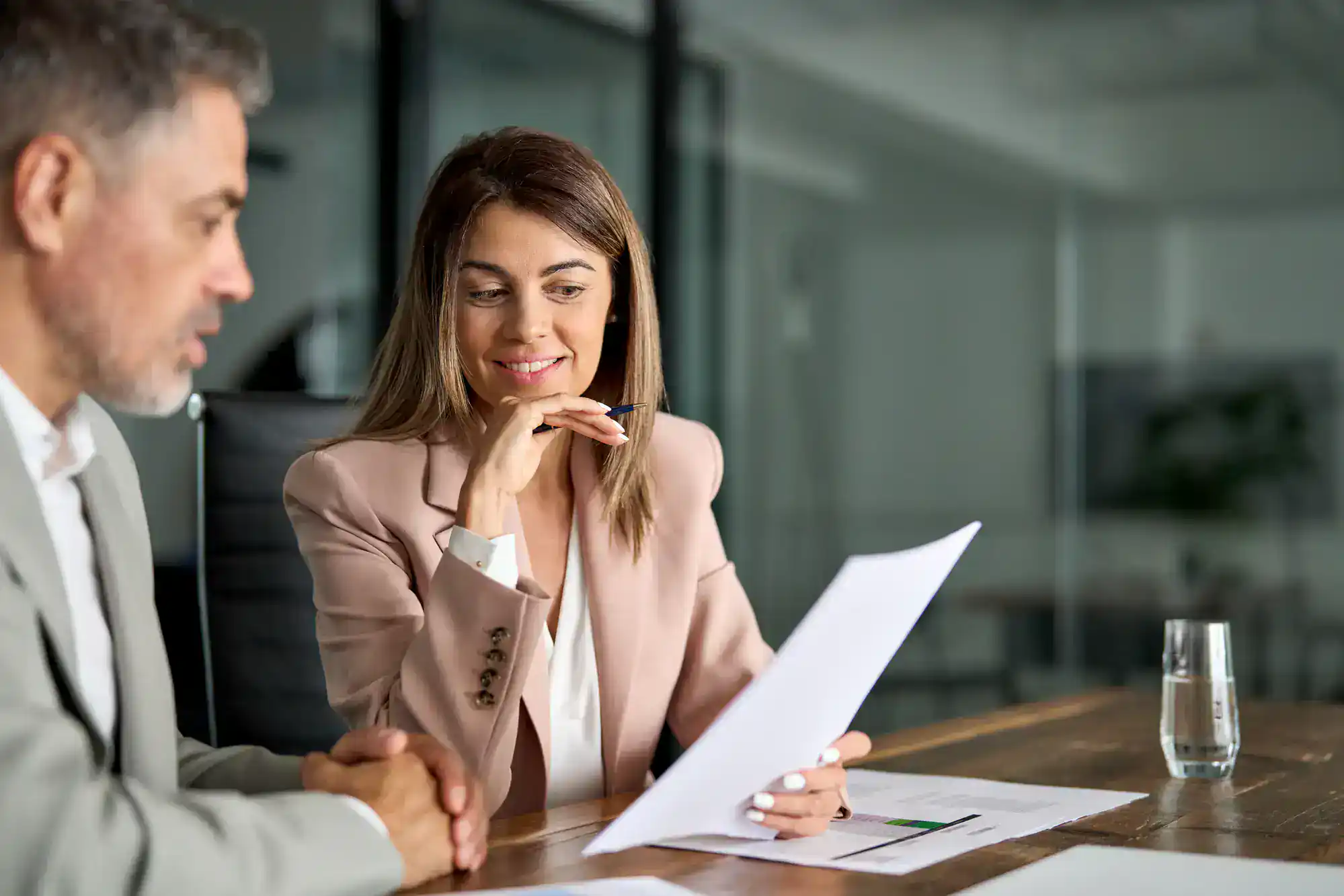 A woman in a light blazer holds and reviews a document with a man in an office setting. Both are seated at a wooden table with papers and a glass of water, appearing engaged in a business discussion.