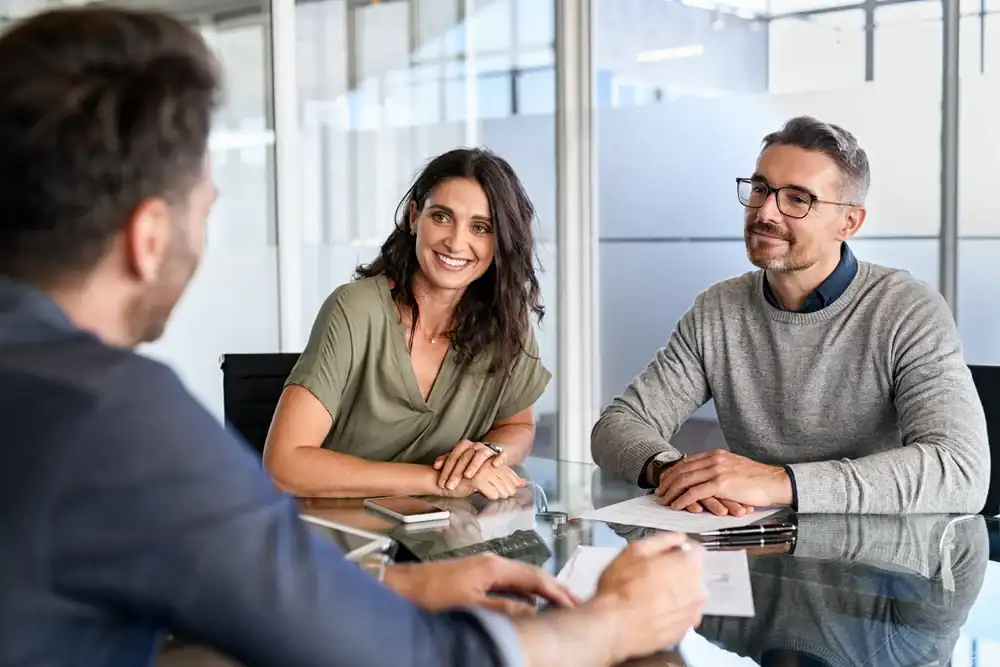 Three people sit at a glass table in an office, having a discussion. A woman and a man face another man whose back is to the camera. The woman is smiling and the atmosphere appears friendly and professional.