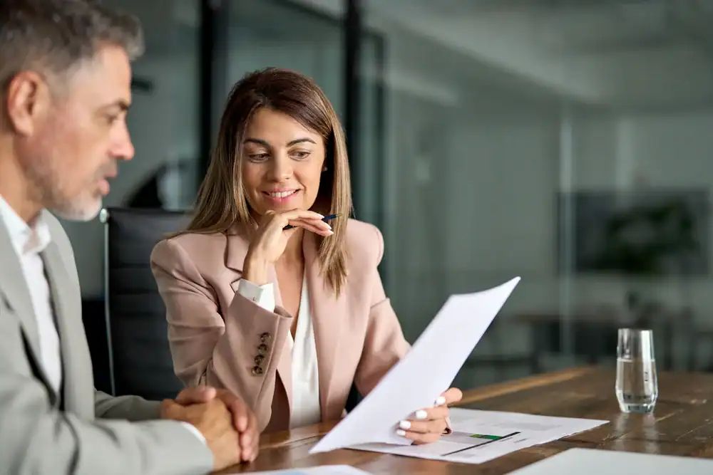 A woman in a light-colored blazer reviews documents with a man in an office setting. She is smiling and holding a pen, while the man listens attentively. A glass of water sits on the table.