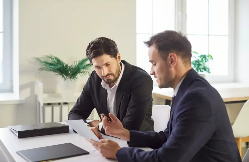 Two men in business suits sit at a desk in a bright office, discussing a document one of them is holding. A closed folder and a pen are on the table, with green plants in the background.